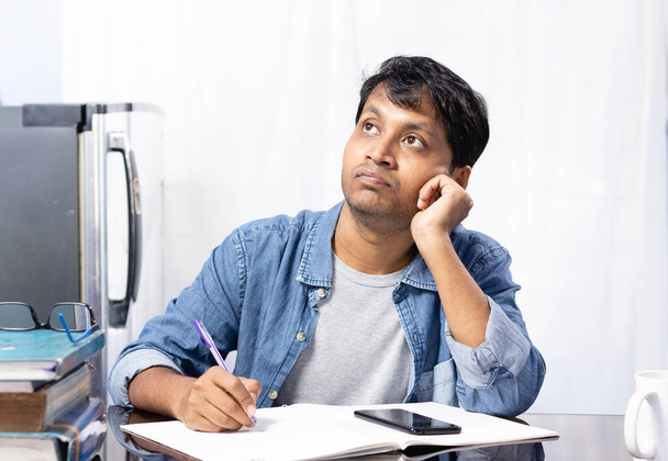 Ein junger indischer Mann blickt nach oben und denkt nach, während er zu Hause auf weißem Hintergrund studiert - Foto, Bild