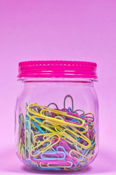 Colorful paperclips in a plain glass jar and sealed lid with pink background and copy space above, creative concept. - Foto, Bild