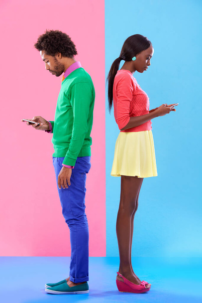 Slightly distracted. Studio shot of a young couple using cellphoes standing against a colourful background - Foto, imagen