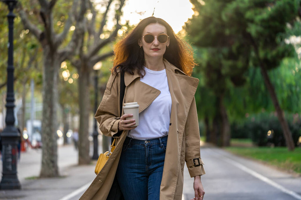 Adult 35s years old businesswoman with long hair, sunglasses and trench coat walk from office to a town with coffee cup. Urban scene of Barcelona city. - Foto, imagen