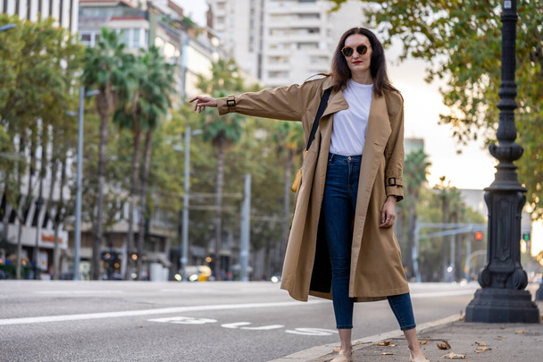 Ενηλίκων 35 ετών επιχειρηματίας με μακριά μαλλιά, γυαλιά ηλίου και καμπαρντίνα πάρετε ένα ταξί στο κέντρο της πόλης με σύγχρονα κτίρια. Αστική σκηνή της πόλης της Βαρκελώνης. - Φωτογραφία, εικόνα