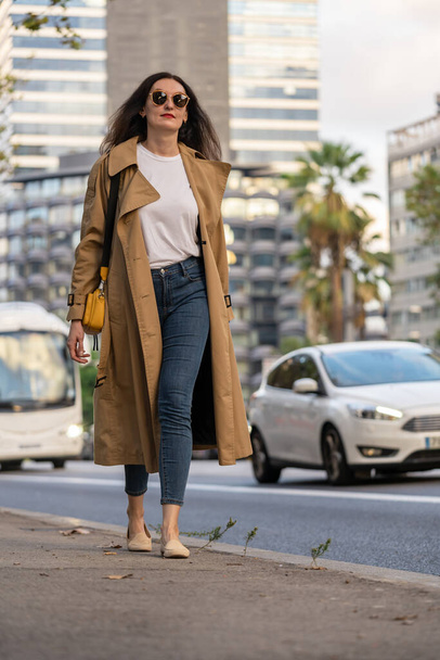 Доросла 35-річна бізнес-леді з довгим волоссям, сонцезахисними окулярами і траншеєю ходять в місті з сучасними будівлями. Міська сцена Барселони. - Фото, зображення