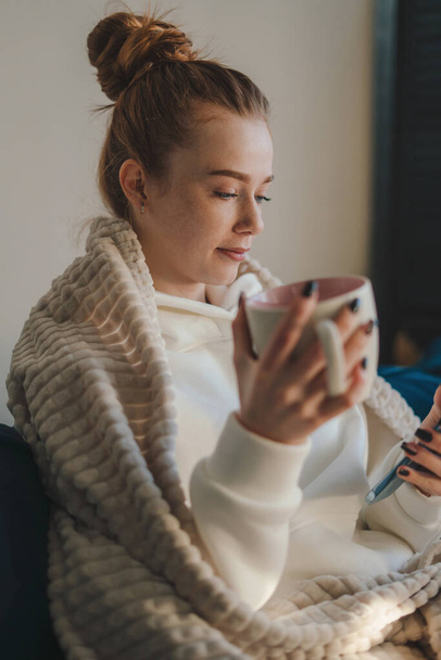 Young beautiful woman sitting on the sofa with cup of hot coffee, feeling happy and smile using smartphone. Internet technology. Internet communication. Cozy - Foto, Imagen