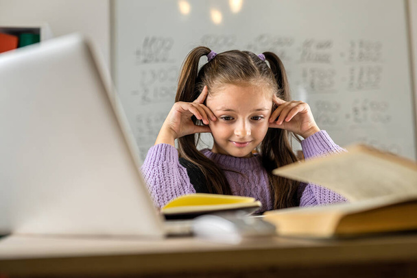 Милая маленькая девочка, использующая ноутбук, чтобы учиться дома. симпатичный ребенок сидит за столом, смотрит на экран компьютера и улыбается. наслаждаясь дистанционным обучением. close up - Фото, изображение