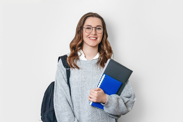 studentka oblečená ve svetru s batohem a brýlemi se usmívá a drží zápisník, sešit, složku na bílém pozadí - Fotografie, Obrázek