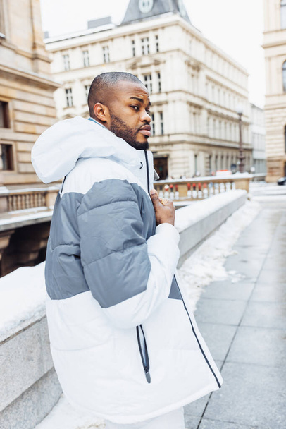 Young African man in casual white and blue clothes, striped sweater. Winter season street fashion portrait with snow. Lifestyle, city life concept. View over shoulder from behind - Fotografie, Obrázek