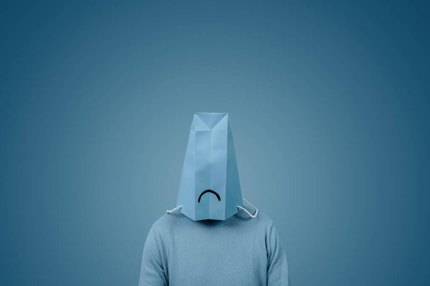男は青い紙の袋を頭の中に持っていて、周りに空白がある青い背景に悲しい口が描かれていました - 写真・画像