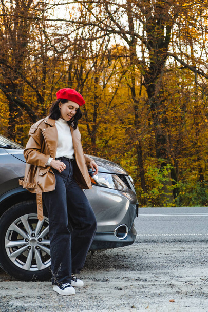 mujer joven en boina roja se encuentra cerca de coche en el fondo del bosque de otoño - Foto, Imagen