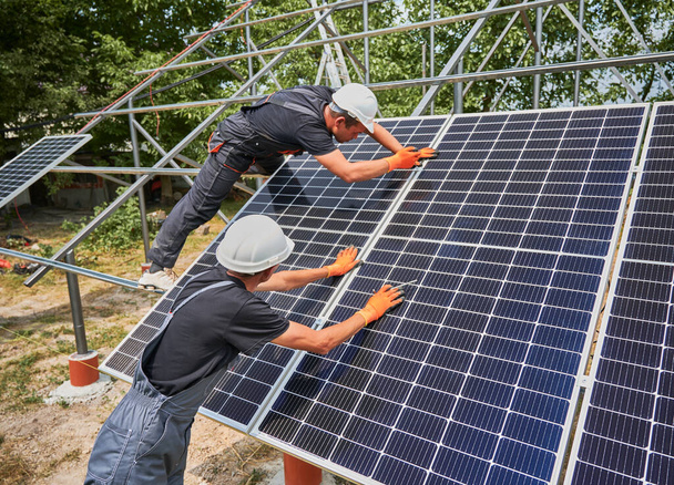 Dışarıya fotovoltaik güneş paneli kuran işçiler. Erkek mühendisler güneş modüllerini metal raylara yerleştiriyor, inşaat kaskları ve iş eldivenleri takıyorlar. Yenilenebilir ve ekolojik enerji. - Fotoğraf, Görsel