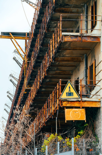 A yellow triangular sign hangs on the dilapidated rusty balconies of an abandoned old multistory building with a large number of lifting mechanisms on the roof. Vertical photo - Fotó, kép