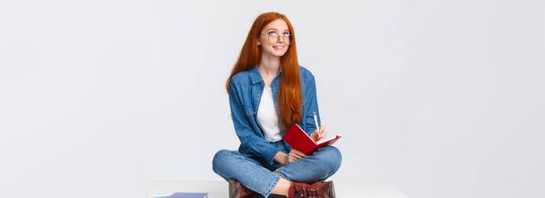 夢のような、創造的でかわいい勤勉な赤い髪の女性の学生は、眼鏡で、足で床に何かを書くノートブックに囲まれて座って、詩を作成し、思慮深い検索. - 写真・画像