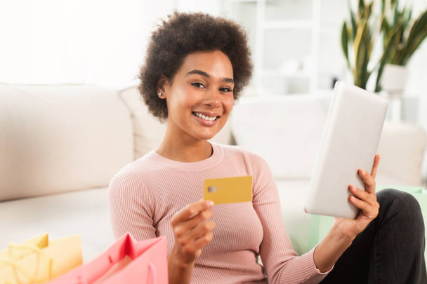 Glad happy young mixed race lady shopaholic with tablet and credit card checking financial bank account in living room interior. Online shopping at home, sale and cashback, app for pay money for order - Photo, Image