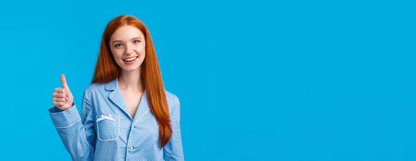 Girl approves recommend product. Cheerful satisfied pretty redhead woman in pyjama, ready sleep, confirm info, showing yes gesture thumbs-up and smiling, standing blue background. - Photo, Image