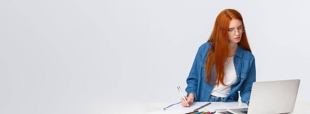 Inspiráció, munka és művészet koncepció. Összpontosított elfoglalt aranyos vörös hajú nő szemüvegben, jegyzeteket ír le a laptopról, hasznos weboldal, online tanfolyam tanulni rajz, fehér háttér. - Fotó, kép