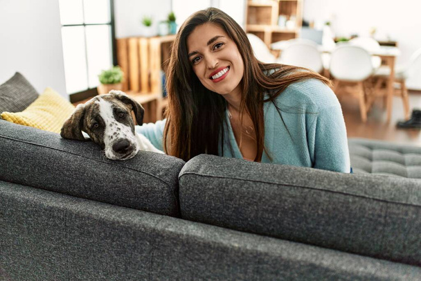 Mujer joven sonriendo confiada sentada en el sofá con el perro en casa - Foto, imagen