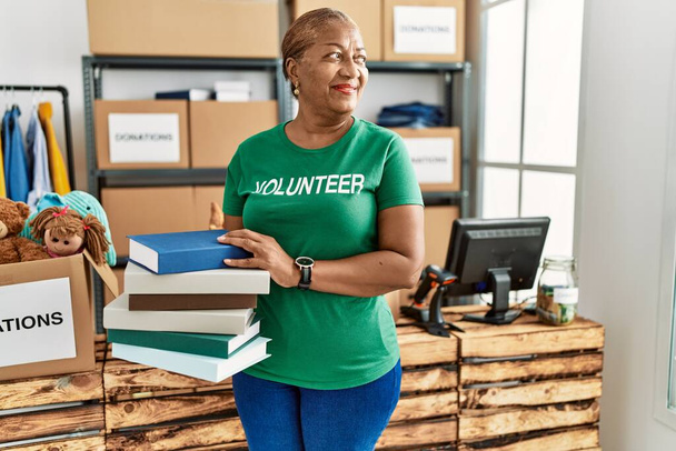 Senior african american woman wearing volunteer uniform holding books at charity center - Photo, Image