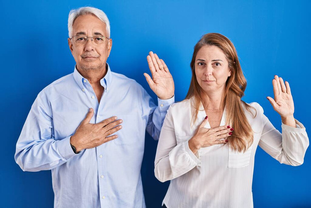Middle age hispanic couple standing over blue background swearing with hand on chest and open palm, making a loyalty promise oath  - Foto, immagini