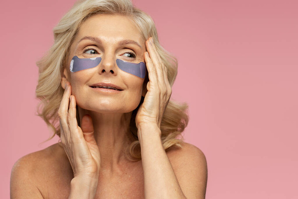 Eye Skin Treatment. Caucasian 50-55 years old blonde mature woman with under collagen smoothing under eye patches on face, looking aside, posing shirtless, isolated over pink background. Copy ad space - Photo, Image