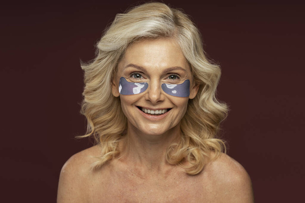 Beautiful Caucasian mature woman with hydrogel collagen eye patches, smoothing dark circles and puffiness under the eyes smiling looking at camera, posing shirtless on color background. Skin care - Photo, Image