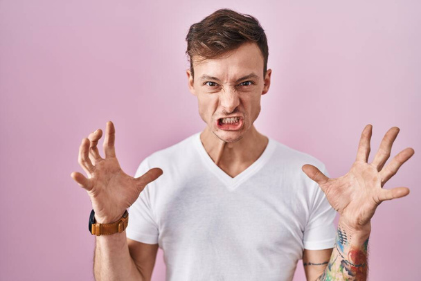 Caucasian man standing over pink background crazy and mad shouting and yelling with aggressive expression and arms raised. frustration concept.  - 写真・画像