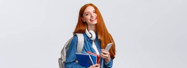 Femme rousse gaie, amicale et charismatique riant joyeusement, envoyant des SMS à des amis, bavardant avec un smartphone, profitant du mode de vie universitaire, se dirigeant vers le campus avec un sac à dos et des cahiers
. - Photo, image