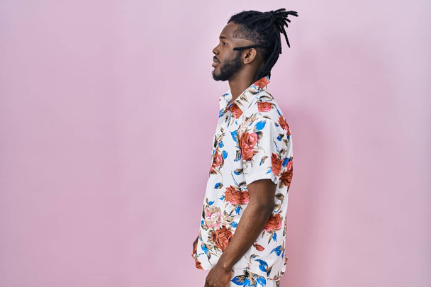 Afrykanin z dredami w letniej koszuli na różowym tle patrząc na bok, relaks profil pozować z naturalną twarzą z pewnym uśmiechem.  - Zdjęcie, obraz