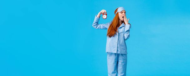 Its so late time to bed. Tired and sleepy cute redhead female in nightwear and sleep mask, holding red alarm clock, yawning turn away with closed eyes, getting ready to sleep, blue background. - Photo, Image