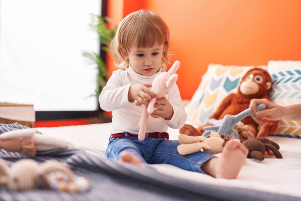 Adorable hispanic girl holding dolls sitting on bed at bedroom - Foto, afbeelding