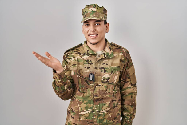 Young arab man wearing camouflage army uniform smiling cheerful presenting and pointing with palm of hand looking at the camera.  - Zdjęcie, obraz