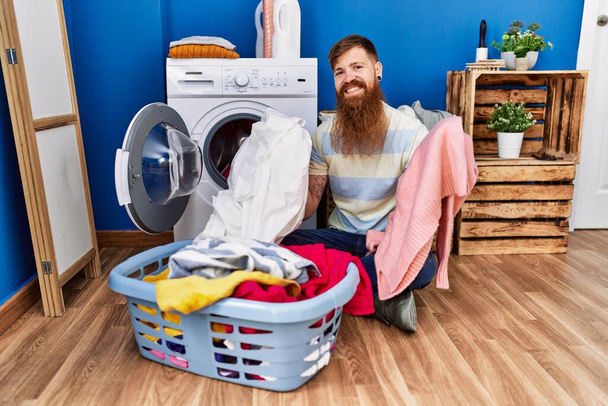 Jeune rousse homme souriant lavage confiant vêtements à la buanderie - Photo, image