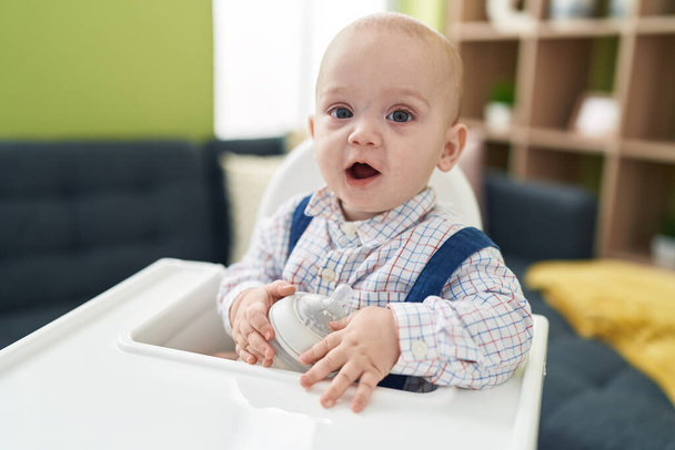 Adorable caucasian baby holding feeding bottle sitting on highchair at home - Photo, Image