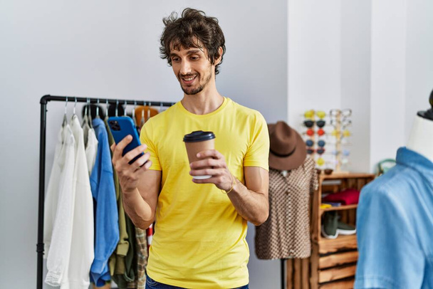 Joven hispano usando smartphone bebiendo café en tienda de ropa - Foto, imagen