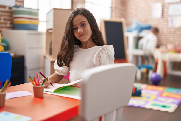 Adorable hispanic girl student sitting on table drawing on paper at kindergarten - Foto, Imagen