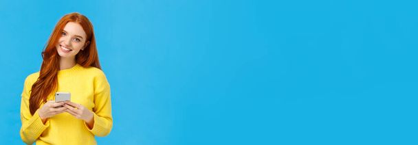 Технологии, интернет и гаджеты. Милая рыжеволосая женщина, отправляющая смс-сообщения, общающаяся с помощью приложения для смартфона, улыбающаяся камера, скачивающая полезное приложение, синий фон
. - Фото, изображение
