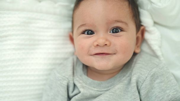 Schattige Spaanse baby glimlachend zelfverzekerd liggend op bed in de slaapkamer - Foto, afbeelding