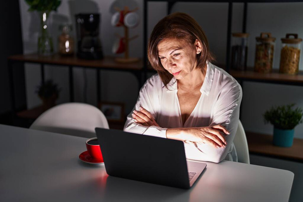 夜間に自宅でノートパソコンを使用している中年のパニック女性は懐疑的で緊張し、両腕を組んで顔に不承認の表現。否定的な人.  - 写真・画像