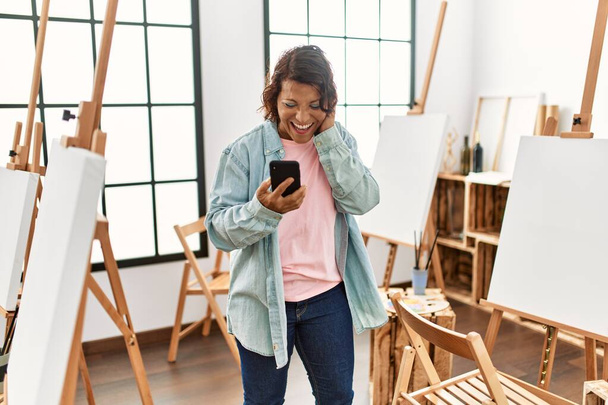 Latynoska artystka w średnim wieku uśmiechnięta szczęśliwa z połączenia wideo za pomocą smartfona w studiu artystycznym. - Zdjęcie, obraz
