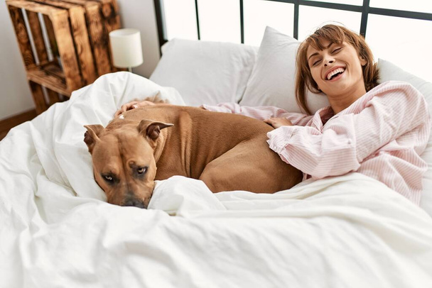 Молодая белая женщина, уверенно улыбающаяся, лежа в постели с собакой в спальне - Фото, изображение
