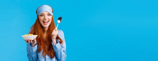 Kävisikö aamiainen? Iloinen hyvännäköinen punapää nainen, jolla on innokas ilme, hymyilevä syöminen viljaa ja lusikka, sininen tausta yöpuvussa, uninaamio. - Valokuva, kuva
