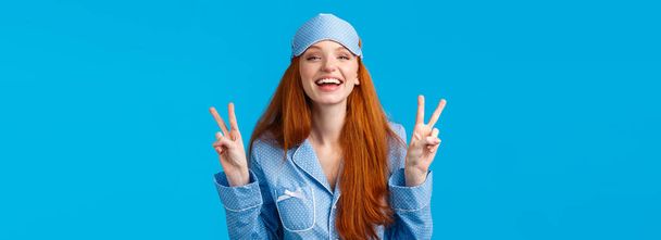Happy and carefree feminine young redhead female in nightwear, sleep mask and pyjama, laughing joyfully, feeling self-assured and pretty showing peace signs, smiling toothy, blue background. - Photo, Image