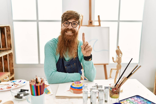 Redhead man with long beard painting clay bowl at art studio showing and pointing up with finger number one while smiling confident and happy.  - Fotó, kép
