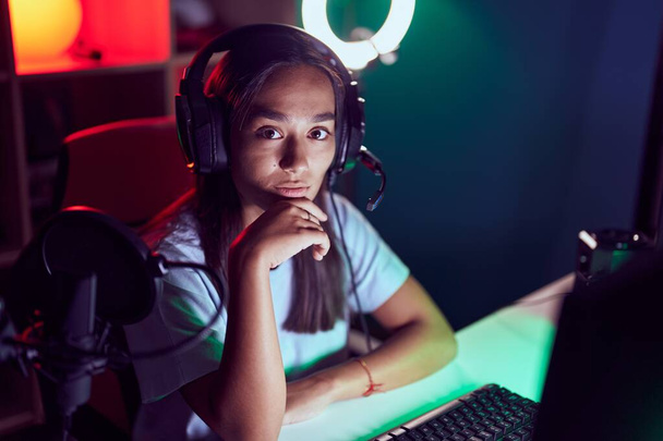 Young beautiful hispanic woman streamer sitting on table with serious expression at gaming room - Zdjęcie, obraz