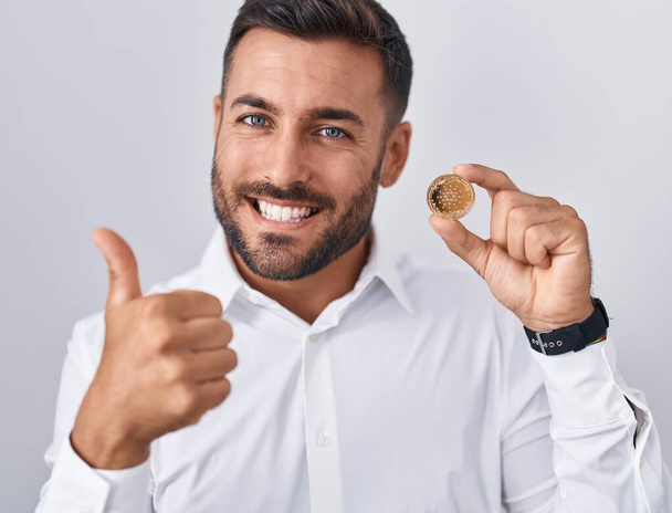 Handsome hispanic man holding cardano cryptocurrency coin smiling happy and positive, thumb up doing excellent and approval sign  - Foto, afbeelding