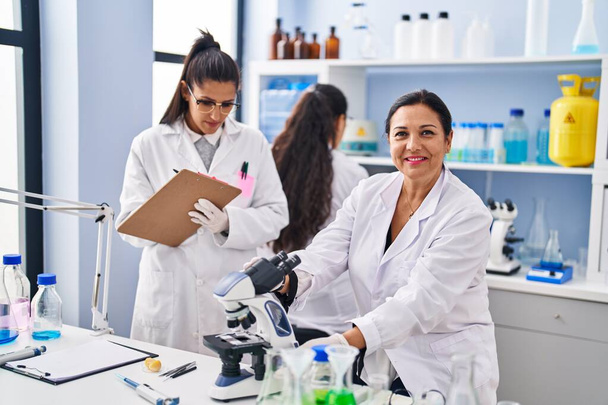 Three woman scientists using microscope write on checklist at laboratory - Photo, image