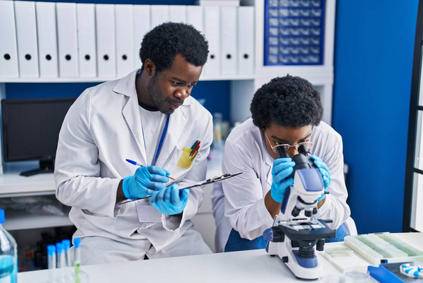 African american man and woman scientists using microscope writing on document at laboratory - Foto, Imagen