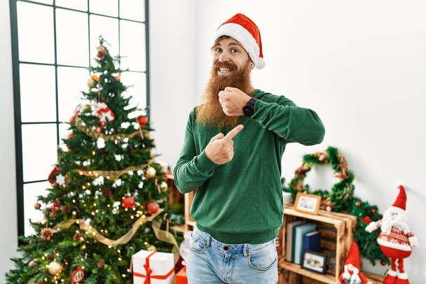 Redhead man with long beard wearing christmas hat by christmas tree in hurry pointing to watch time, impatience, looking at the camera with relaxed expression  - Foto, Bild