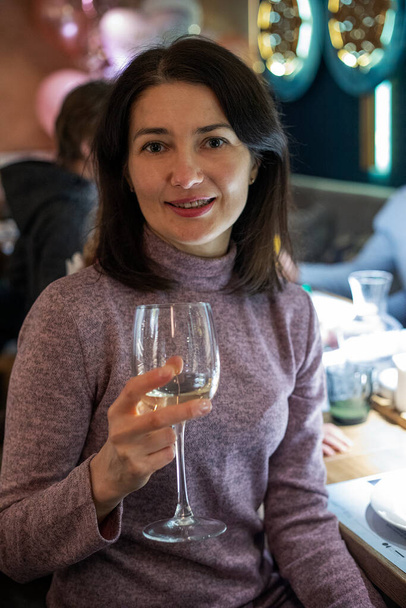 gelukkig brunette meisje in een lila trui voor haar verjaardag met een glas witte wijn. Een feestje. - Foto, afbeelding