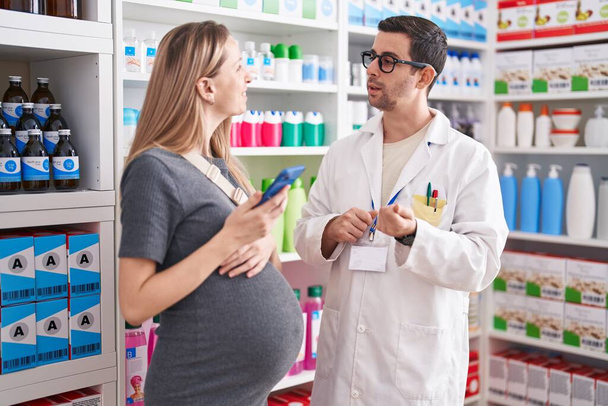 Man and woman pharmacist and pregnant client using smartphone speaking at pharmacy - Фото, изображение