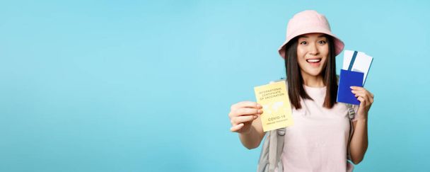 Энтузиастский азиатский турист, девушка показывает ковид-19 международный сертификат прививки, паспорт с двумя билетами, путешествия после коронавирусной вакцины, синий фон. - Фото, изображение