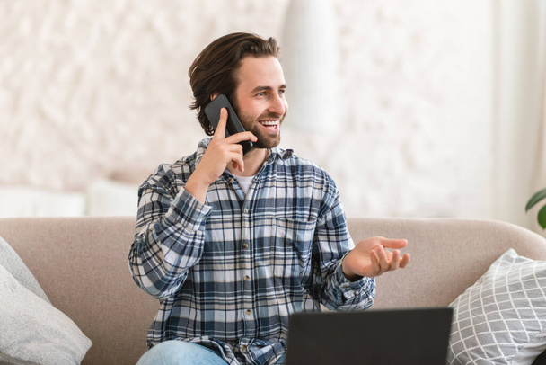 Cheerful busy young caucasian male manager sits on sofa with computer, calls by phone, talks with client, looks at free space in white room interior. Business, work remotely at home, ad and offer - Foto, Imagen